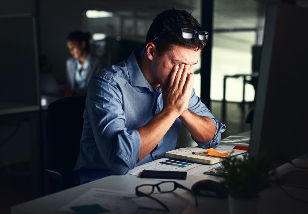 Stressed man working late in a dark office, showing signs of mental fatigue and headache related to business deadlines, with potential links to stress, elevated blood sugar, and diabetes risk.