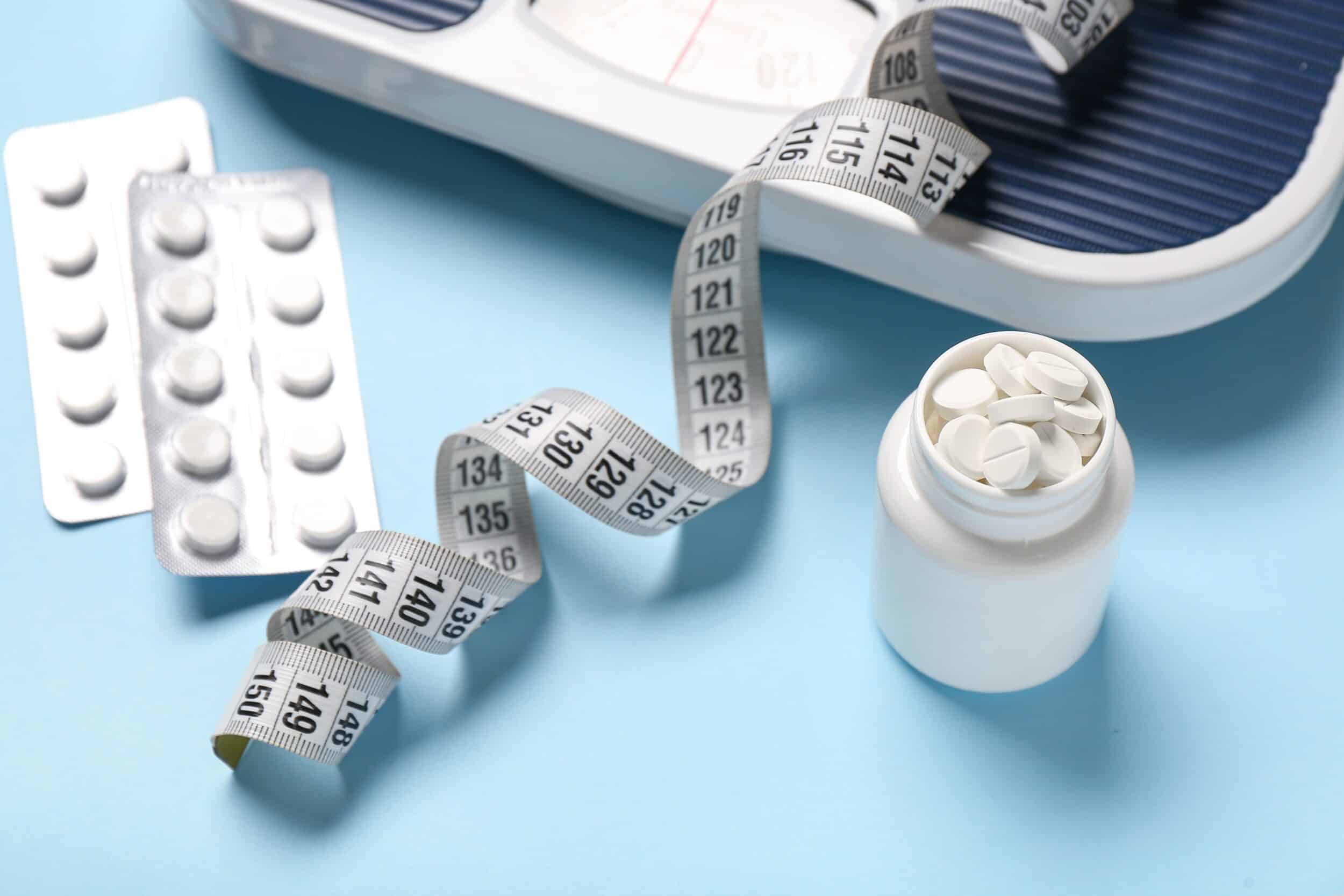 Bottle of weight loss pills next to a measuring tape and bathroom scale on blue background, representing medical weight loss support and treatment options